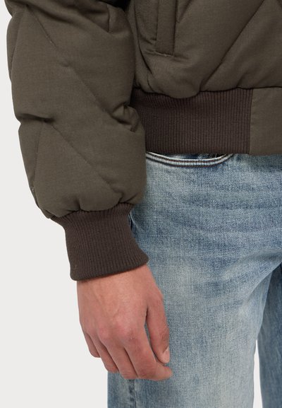 Sötétzöld pufferszákó, steppelt kialakítással, bordó bordázott mandzsettákkal és laza fazonnal. Fénykék farmernadrággal párosítva.