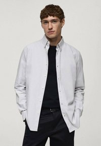 Cămașă butonată de culoare gri deschis, cu guler clasic, mâneci lungi și o textură subtilă, purtată peste un tricou negru cu guler rotund și pantaloni închiși la culoare.