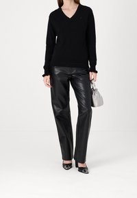 Pull noir à col en V, manches longues avec des poignets à volants, pantalon en cuir noir droit, sac à main gris et chaussures à talons noires avec accents brillants.