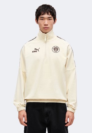 Jeune homme portant un sweat-shirt crème à fermeture éclair partielle avec les logos FC St. Pauli et Puma, associé à un pantalon noir, debout devant un fond uni.