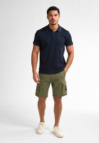 Navy polo shirt with a collar and striped accents, paired with olive green cargo shorts featuring side pockets and patches, and beige sneakers.