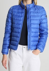 Blå puffjacka i glänsande material, med hög krage, framdragkedja, vertikal quiltning och en rund logo på ärmen.