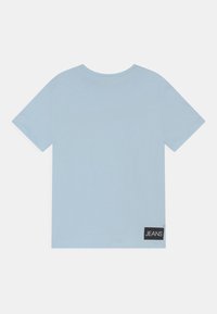 Hellblaues T-Shirt mit kurzen Ärmeln, rundem Halsausschnitt und einem schwarzen Etikett, auf dem "JEANS" im unteren rechten Bereich steht. Glatte Stoffstruktur.