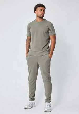 Homme avec des tatouages portant un t-shirt vert olive et un pantalon, debout les mains dans les poches, regardant légèrement sur le côté, avec des baskets blanches aux pieds.