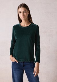 Haut long en velours vert avec un col rond et une surface texturée. Présente un accent à cordon au niveau de l'ourlet. Associé à un jean en denim foncé.