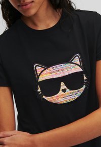 T-shirt nera con un colorato volto di gatto ricamato che indossa occhiali da sole. Il design presenta strisce multicolori e una texture liscia.