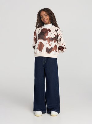 Ragazza con capelli ricci che indossa una felpa marrone e bianca tie-dye, jeans a gamba larga scuri e sneakers bianche, in piedi con le mani dietro la schiena.