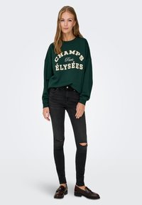 Felpa verde con testo bianco "CHAMPS ÉLYSÉES", abbinata a jeans skinny neri strappati e mocassini scuri, su uno sfondo neutro.