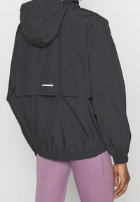 Černá lehká bunda s kapucí, elastickým pasem a dlouhými rukávy. Na zádech má reflexní prvek. Hladká textura.