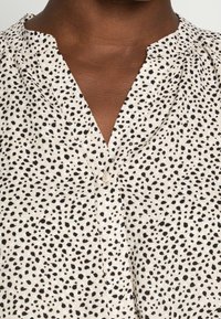 Hellbeige Bluse mit schwarzem Punktemuster, Knopfverschluss und weicher Textur. Verfügt über einen V-Ausschnitt und dezente Raffung an den Schultern.