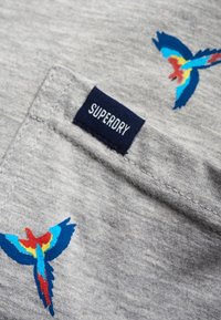 Šedá textilie s barevnou výšivkou ptáka v červené, modré a žluté barvě. Obsahuje námořnický štítek s nápisem "SUPERDRY" na švu.