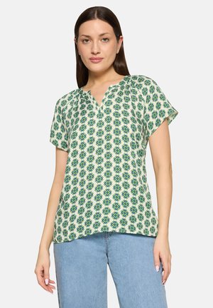 Frau mit glatten dunklen Haaren trägt ein weißes Kurzarmshirt mit blau-grünem Blumenmuster und hellblauen Jeans, steht vor neutralem Hintergrund.