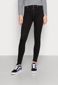 Czarne, wysmukłe dżinsy z wysokim stanem i postrzępionym wykończeniem, zestawione z czarno-niebieskimi sneakersami w wzory. Sweter ma teksturowany, szary splot.