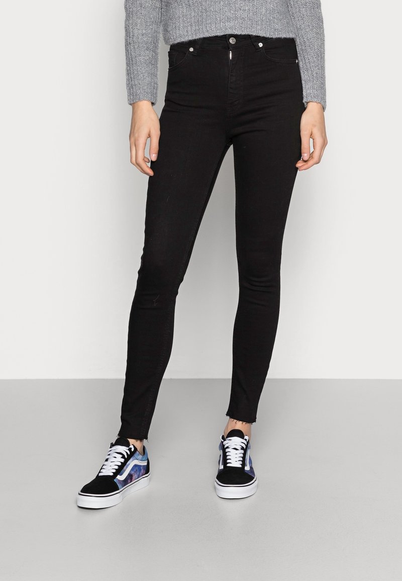 Czarne, wysmukłe dżinsy z wysokim stanem i postrzępionym wykończeniem, zestawione z czarno-niebieskimi sneakersami w wzory. Sweter ma teksturowany, szary splot.