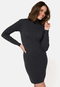 Femme dans une robe tricotée grise foncée, à manches longues et longueur genoux, souriant avec une main sur la hanche et l'autre près de son visage.