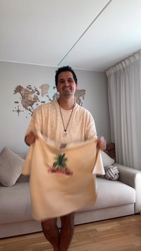 Beige t-shirt met een levendige palmboomprint; gestructureerde stof, losse pasvorm, gedragen door een individu in een woonkamer.