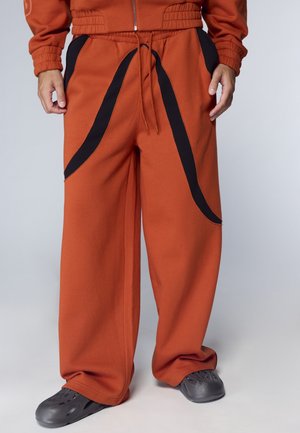 Persona che indossa pantaloni arancioni larghi con strisce curve nere e scarpe slip-on nere, in piedi su uno sfondo chiaro.