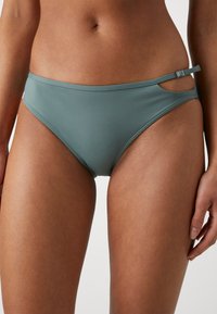 Šviesiai žalia bikini kelnaitė, pagaminta iš lygaus medžiagos, su išpjovos dizainu ir reguliuojamomis šoninėmis juostomis su užsegimu.