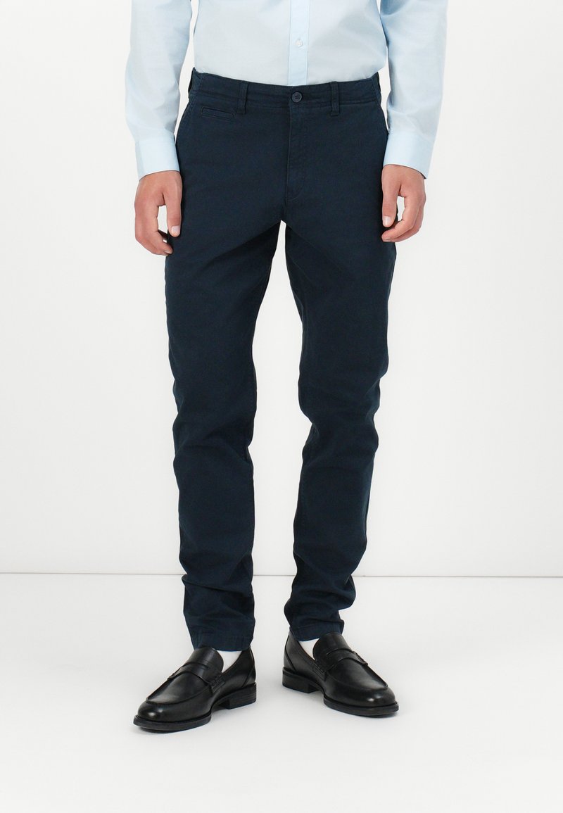 Marineblaue Chinos aus Baumwolle mit Slim-Fit, ausgestattet mit einem Knopfverschluss und Seitentaschen. Kombiniert mit schwarzen Loafers und weißen Socken.