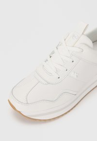 Zapatilla de cuero blanco con puntera redondeada, cierre con cordones y detalles de costura sutiles. Suela de goma con un logo acentuado en marrón en el lateral.