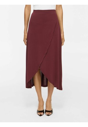 Femme portant une jupe midi taille haute style portefeuille rouge foncé associée à des talons ouverts noirs sur un fond uni.
