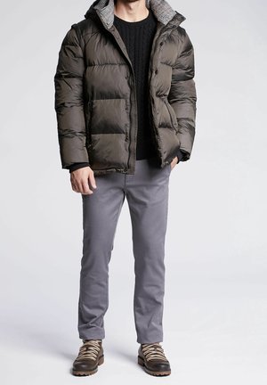 Homme portant une doudoune sombre, un pull en laine noir, un pantalon gris et des bottines marron à lacets, debout devant un fond uni.