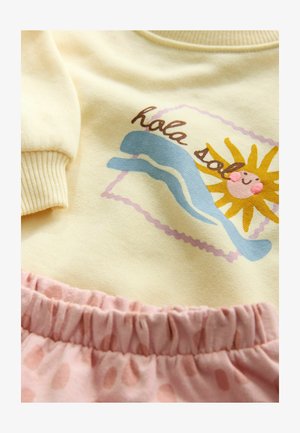 Sárga pamut pulóver napsütés és hullám grafikával, rózsaszín szoknyával párosítva, amely finom pöttyös mintával és rugalmas derékpánttal rendelkezik.