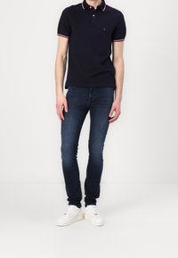 Man med en marinblå polo med vit och röd kant, smala mörkblå jeans och vita sneakers, stående mot en enkel vit bakgrund.