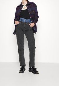 Jeans negros de corte recto, combinados con una camisa de cuadros en púrpura y verde y zapatos de charol negros. La camisa tiene botones dorados.