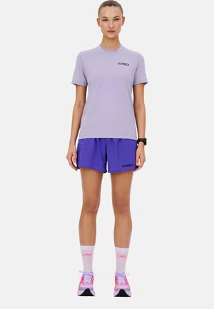 Lila Sport-T-Shirt mit schwarzem Logo, kombiniert mit lila Shorts. Weiße Socken mit pinkem Logo. Die Schuhe sind pink-graue Laufschuhe.
