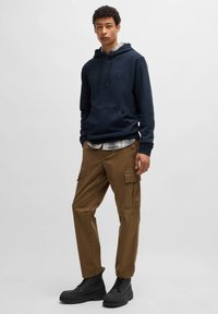 Navig blauwe hooded sweatshirt met kangoeroezak, gelaagd over een geruit overhemd, gecombineerd met bruine cargobroeken en zwarte veterschoenen.