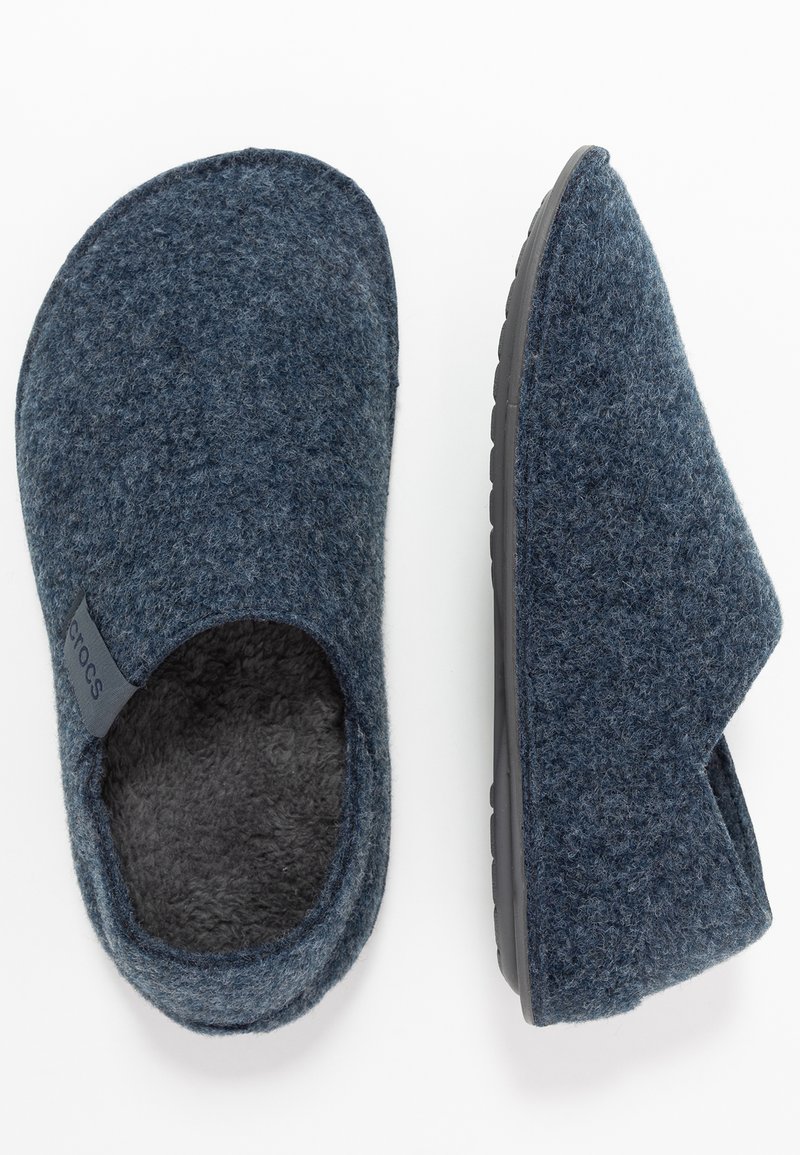 Zapatillas de fieltro azul oscuro con un interior suave, diseño abierto, punta redondeada y suela de goma. Presenta un acento de marca en el lateral.