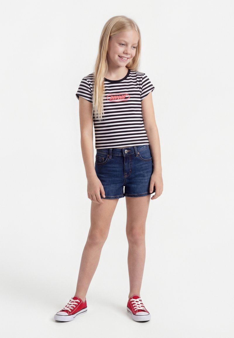 Ξανθιά κοπέλα με χαμόγελο, φοράει κοντομάνικο μαύρο-άσπρο ριγέ μπλουζάκι, σκούρα τζιν σορτς και κόκκινα αθλητικά παπούτσια, στέκεται μπροστά σε λευκό φόντο.