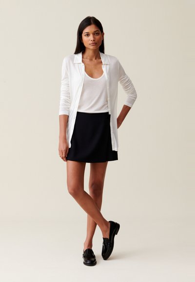 Cardigan blanc à boutons sur un débardeur blanc, associé à une jupe noire et des mocassins noirs ; tissu lisse et éléments de design classiques.