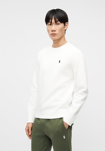 Νέος άνδρας που φοράει λευκό πουκάμισο με μακριά μανίκια και πράσινο παντελόνι με μικρά μαύρα και λευκά λογότυπα polo player, στέκεται μπροστά σε μια απλή φόντο.