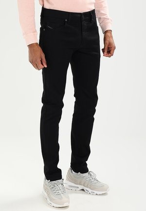 Mann trägt schlanke schwarze Jeans, hellrosa langärmliges Hemd und weiß-graue Turnschuhe, steht vor einem schlichten hellen Hintergrund.