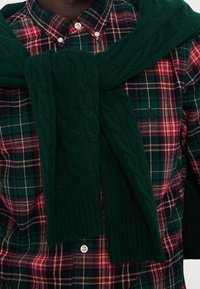 Camicia a quadri in verde scuro, rosso e bianco con un maglione in maglia cable-knit verde scuro annodato al collo; visibile dettaglio del tessuto testurizzato.