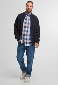 Marineblauer Strickcardigan über einem karierten Hemd in Hellblau, Pink und Weiß, kombiniert mit hochgekrempelten blauen Jeans und beigen Sneakers.