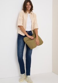 Camisa beige de botones, top blanco, jeans oscuros azules, sosteniendo un bolso acolchado verde oliva, zapatillas crema, de pie contra un fondo liso.