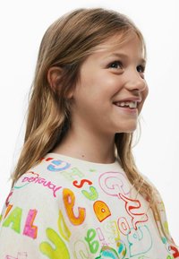 Šviesiai smėlio spalvos megztinis su ryškiomis, didelėmis raidėmis ir abstrakčiomis formomis įvairių spalvų. Minkšta tekstūra ir laisvas kirpimas.