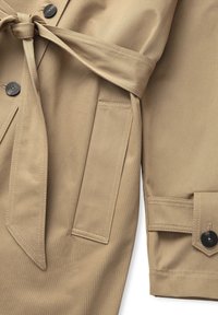 Beige trenčkot s tkanou texturou, páskem v oblasti pasu, velkými knoflíky a dvěma bočními kapsami. Vyznačuje se čistým, přizpůsobeným designem.