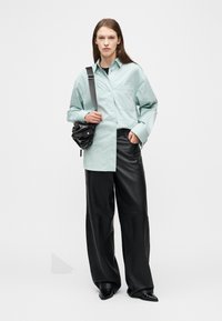 Mujer de pie, vestida con una camisa holgada de color verde azulado claro, pantalones anchos de cuero negros, zapatos negros puntiagudos y llevando un bolso de hombro negro.