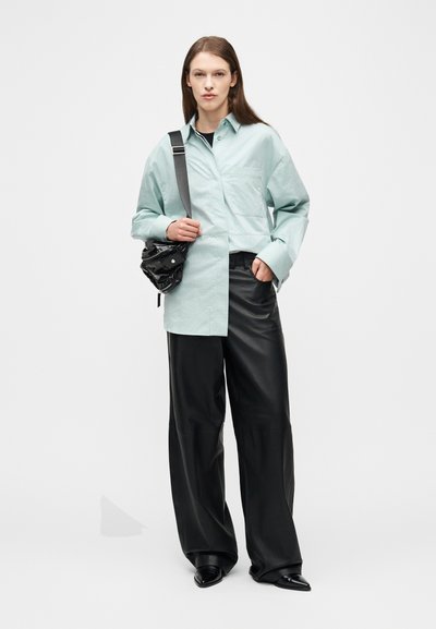 Γυναίκα που στέκεται, φοράει ανοιχτό πράσινο-γαλάζιο φαρδύ πουκάμισο, μαύρο δερμάτινο παντελόνι με φαρδιά πόδια, μαύρα μυτερά παπούτσια και κρατά μια μαύρη τσάντα στον ώμο.