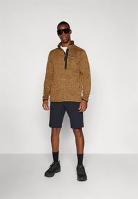 Rjava flis jakna z zadrgo, črni poudarki, kombinirana s temno modrimi hlačami in črnimi čevlji. Model nosi sončna očala in stoji na enostavni podlagi.