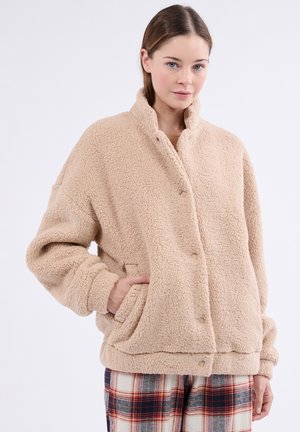 Chaqueta de forro polar beige con una superficie suave y texturizada, cuello alzado, cierres a presión y bolsillos laterales. Combinada con pantalones de patrón de cuadros.