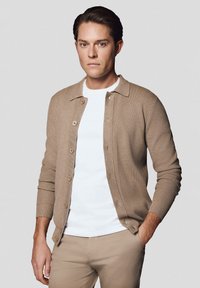 Beżowy kardigan z kołnierzykiem, zapinany na guziki, z ribbottowymi mankietami, noszony na białym T-shircie i beżowych spodniach. Gładka tekstura.