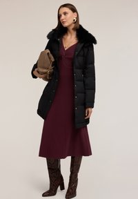 Czarna kurtka puchowa z kołnierzem z sztucznego futra, prążkowana bordowa sukienka i brązowe kozaki do kolan w wężowy wzór. Brązowa torba ze złotymi detalami.