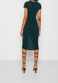 Donna che indossa un abito verde scuro aderente, lungo fino al ginocchio, con maniche corte e sandali con tacco alto neri lacciati, in piedi contro uno sfondo semplice.