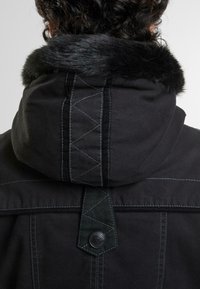 Svart jakke med faux fur-krage, med synlige sømmer, en heksagonal knappdetalj og et trekantet mønster på bakstoppen.
