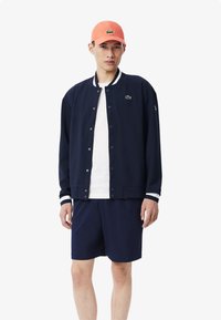 Chaqueta de piloto de la marina con puños y cuello a rayas blancas, camiseta blanca, pantalones cortos azul marino y gorra naranja con logo. Tela suave y ligera.